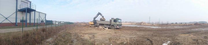 Żółta koparka ładuje ziemię na wywrotkę na placu budowy. W tle budynek z metalową konstrukcją i ogrodzenie z siatki.