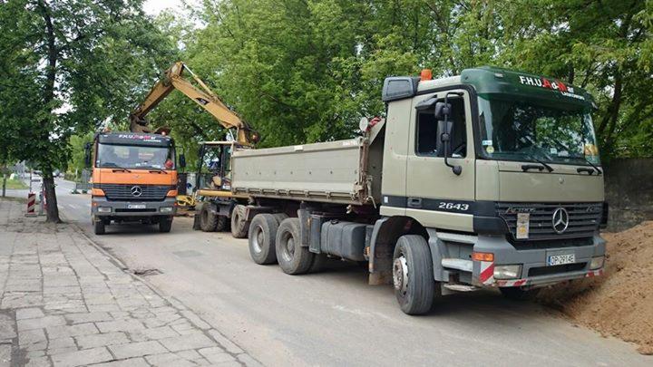 Dwa samochody ciężarowe Mercedes i koparka podczas załadunku ziemi na poboczu drogi w słoneczny dzień. Widoczne drzewa w tle.