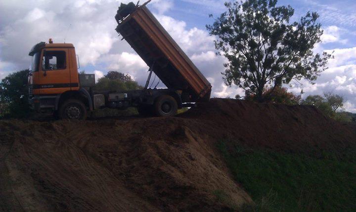 Pomarańczowa wywrotka MAN 18-40 wyładowuje ziemię na nierównym terenie, z drzewem w tle i pochmurnym niebem.