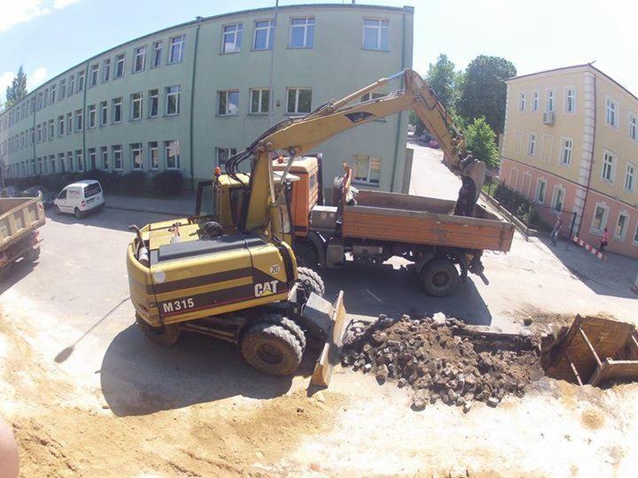 Żółty koparko-ładowarka CAT M315 ładuje ziemię do pomarańczowej wywrotki na tle budynku. Wykopany dół z pryzmą ziemi obok. Ujęcie z góry.