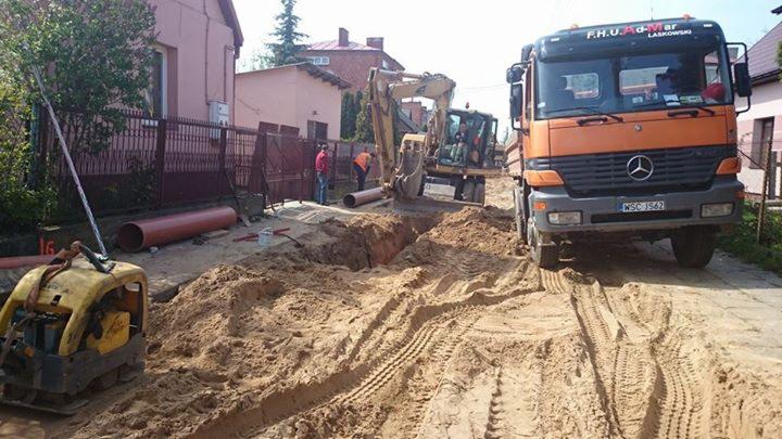 Żółta koparka układa rury w wykopie. Obok stoi pomarańczowa ciężarówka i ubijarka. Pracownicy w odblaskowych kamizelkach nadzorują proces.