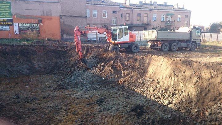 Pomarańczowo-biała koparka Atlas i wywrotka na placu budowy, wykop pod fundamenty. Budynek w tle. Tablice reklamowe widoczne po lewej stronie.