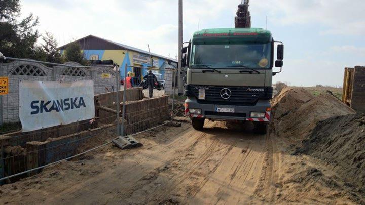 Ciężarówka z dźwigiem na placu budowy Skanska. Widoczne zabezpieczenia wykopu, budynek w tle i pracownicy w odblaskowych kamizelkach. Ujęcie z poziomu gruntu.