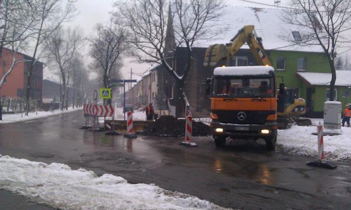 Zimowy krajobraz miejski z wykopem zabezpieczonym znakami drogowymi. Pomarańczowa ciężarówka i koparka CAT w tle. Prace ziemne na mokrej jezdni.