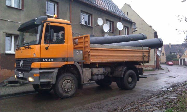 Pomarańczowa ciężarówka Mercedes 1840 przewozi czarne rury na skrzyni ładunkowej, w tle budynki mieszkalne i mokra ulica, pochmurny dzień.