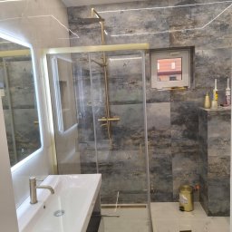 SELBUD Sp. z o.o. - Nowoczesna łazienka z kabiną prysznicową w złotych ramach, umywalką z szafką i podświetlanym lustrem. Płytki w odcieniach szarości i beżu. Oświetlenie LED.