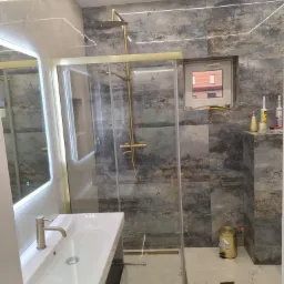 Nowoczesna łazienka z kabiną prysznicową w złotych ramach, umywalką z szafką i podświetlanym lustrem. Płytki w odcieniach szarości i beżu. Oświetlenie LED.