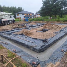 SELBUD Sp. z o.o. - Wylane fundamenty pod dom jednorodzinny, zabezpieczone folią, z betonomieszarką w tle i widokiem na budynki w oddali. Etap budowy domu.