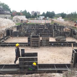 SELBUD Sp. z o.o. - Fundamenty domu w trakcie budowy: zbrojenie, szalunki, pracownicy w kaskach. Na drugim planie domy i materiały budowlane.