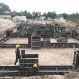 Fundamenty domu w trakcie budowy: zbrojenie, szalunki, pracownicy w kaskach. Na drugim planie domy i materiały budowlane.