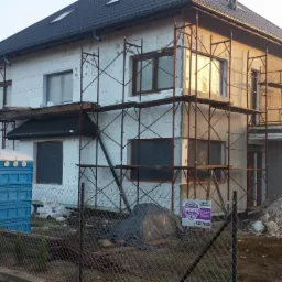 Dwukondygnacyjny dom w trakcie ocieplania, widoczne rusztowania, styropian na ścianach, przenośna toaleta na pierwszym planie i tablica reklamowa firmy ochroniarskiej