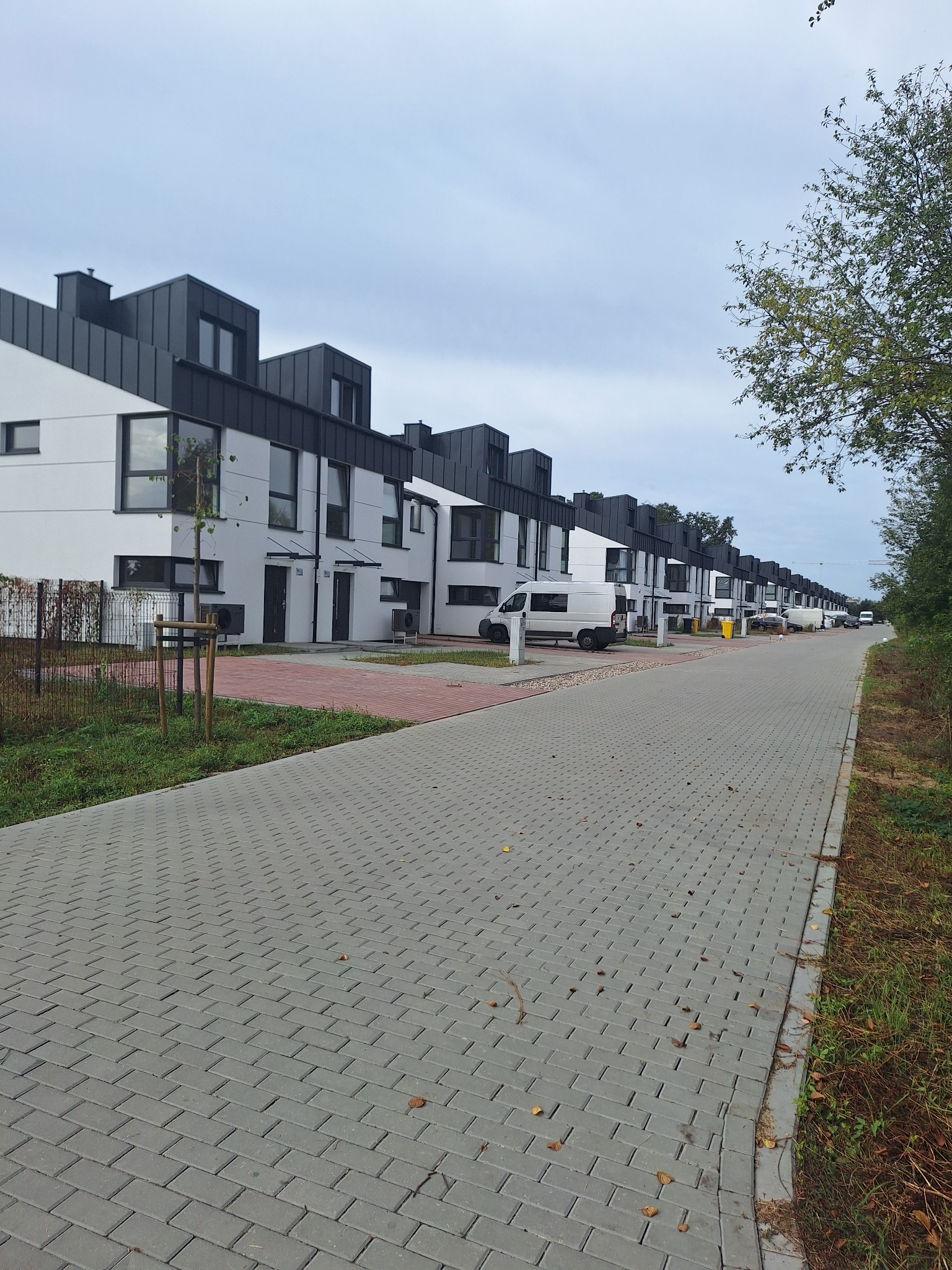 Długi rząd nowoczesnych, szeregowych domów z płaskimi, ciemnymi dachami i białymi elewacjami, widzianych z perspektywy chodnika z kostki brukowej, z zaparkowanymi samochodami dostawczymi w tle,...