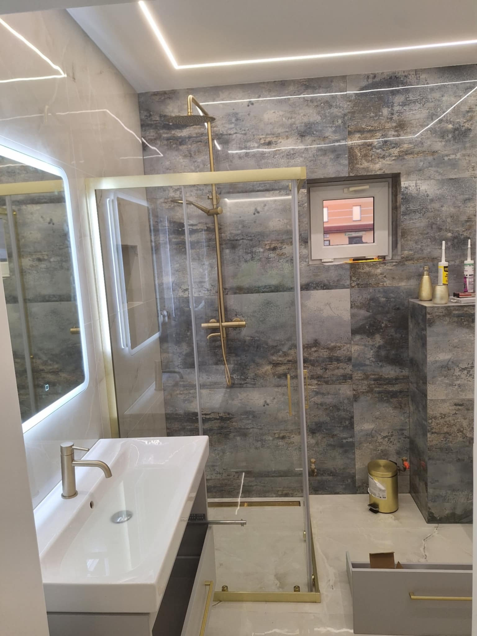 Nowoczesna łazienka z kabiną prysznicową w złotych ramach, umywalką z szafką i podświetlanym lustrem. Płytki w odcieniach szarości i beżu. Oświetlenie LED.