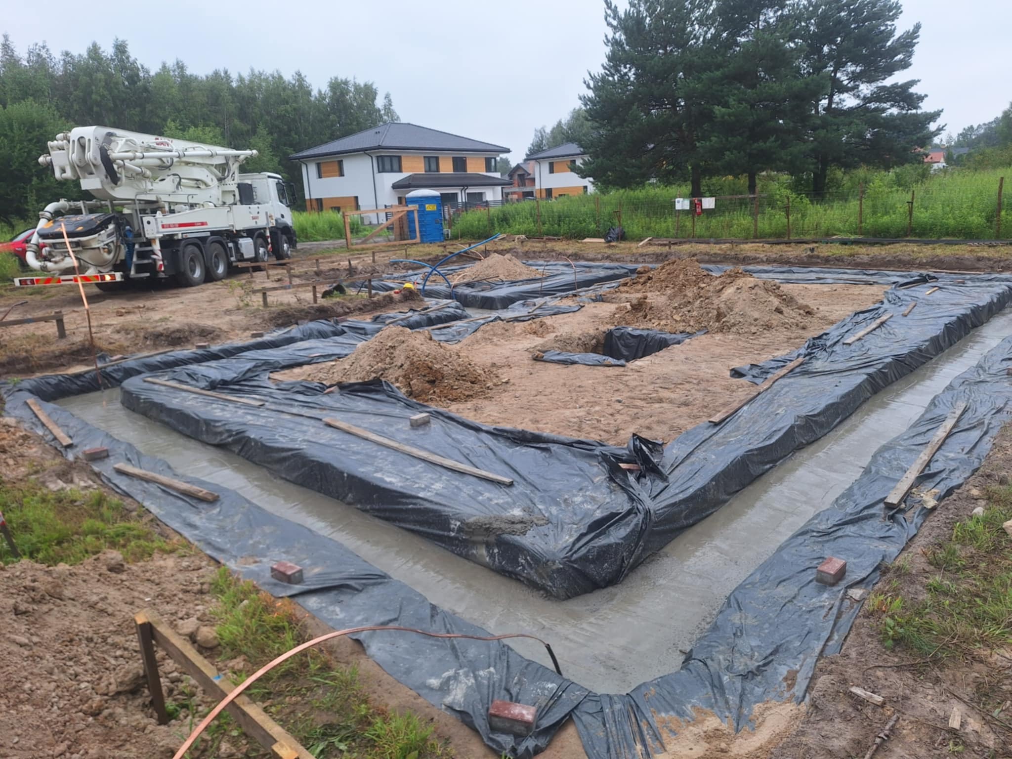 Wylane fundamenty pod dom jednorodzinny, zabezpieczone folią, z betonomieszarką w tle i widokiem na budynki w oddali. Etap budowy domu.
