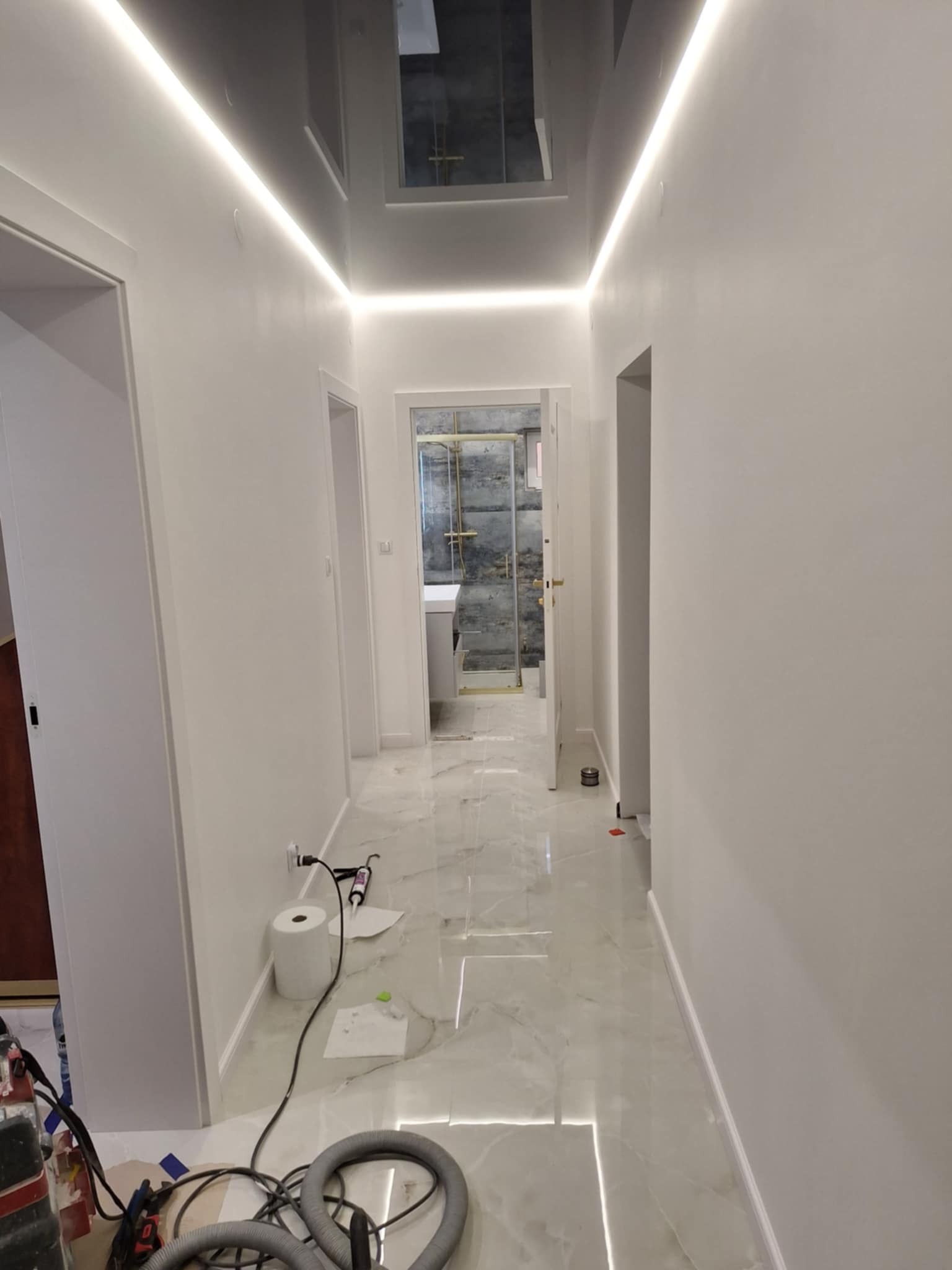Biały korytarz z połyskiem, marmurowa podłoga i ledowe oświetlenie sufitowe. Widoczne narzędzia pracy w rogu, sugerujące trwający remont.
