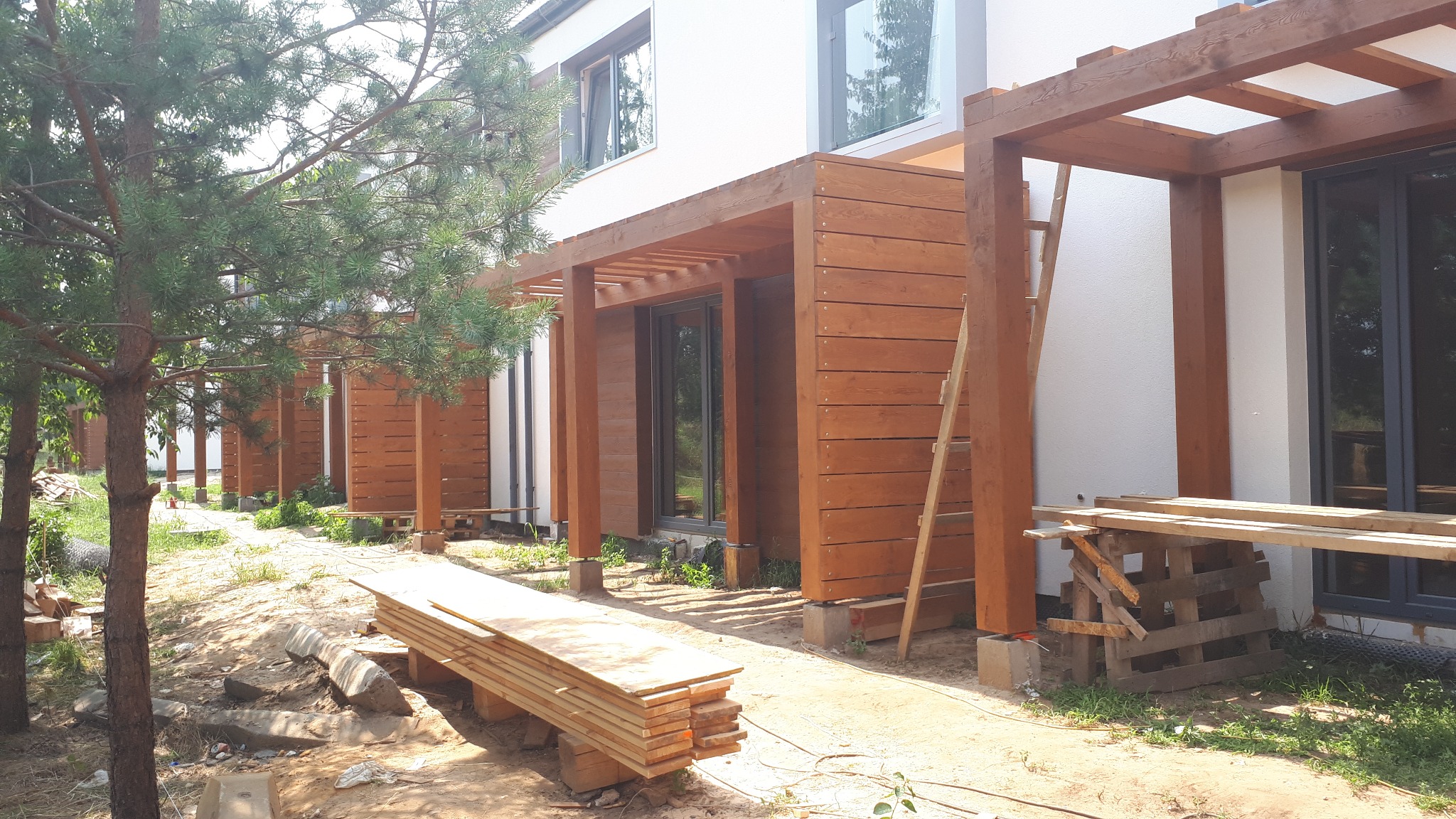 Drewniane pergole przy domu w Błoniu, w trakcie montażu. Widoczne deski, drabina i elementy konstrukcyjne. Prace wykończeniowe elewacji zewnętrznej.