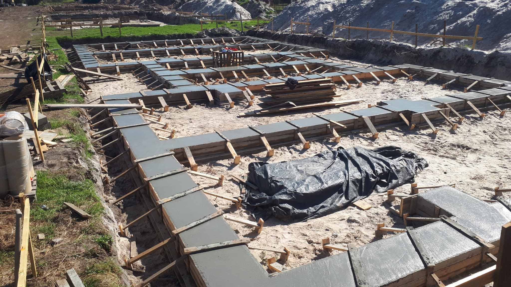 Wylane fundamenty domu z drewnianym szalunkiem, widoczne z góry. Szary beton kontrastuje z piaskowym podłożem. Część fundamentów przykryta czarną folią.