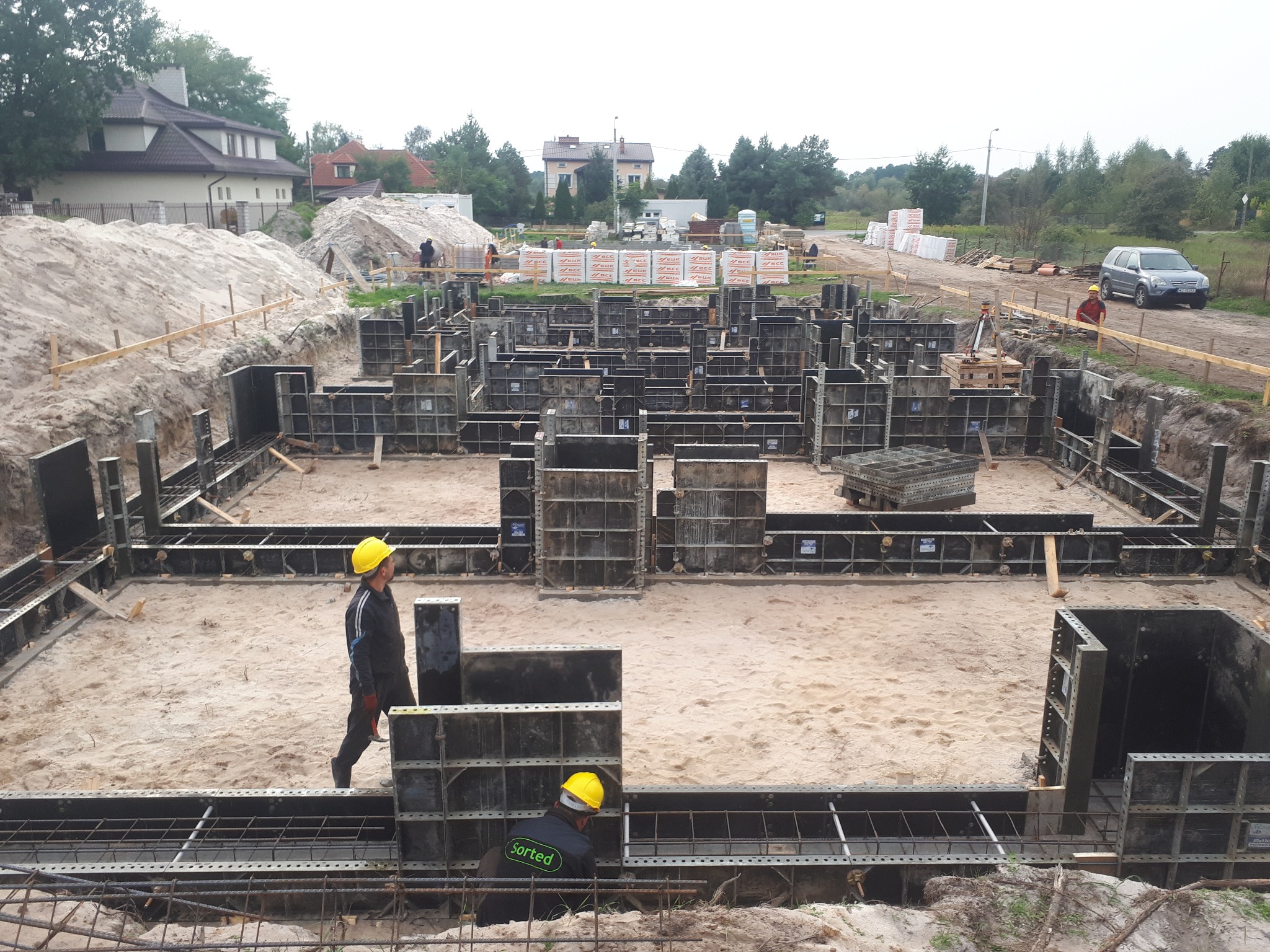 Fundamenty domu w trakcie budowy: zbrojenie, szalunki, pracownicy w kaskach. Na drugim planie domy i materiały budowlane.
