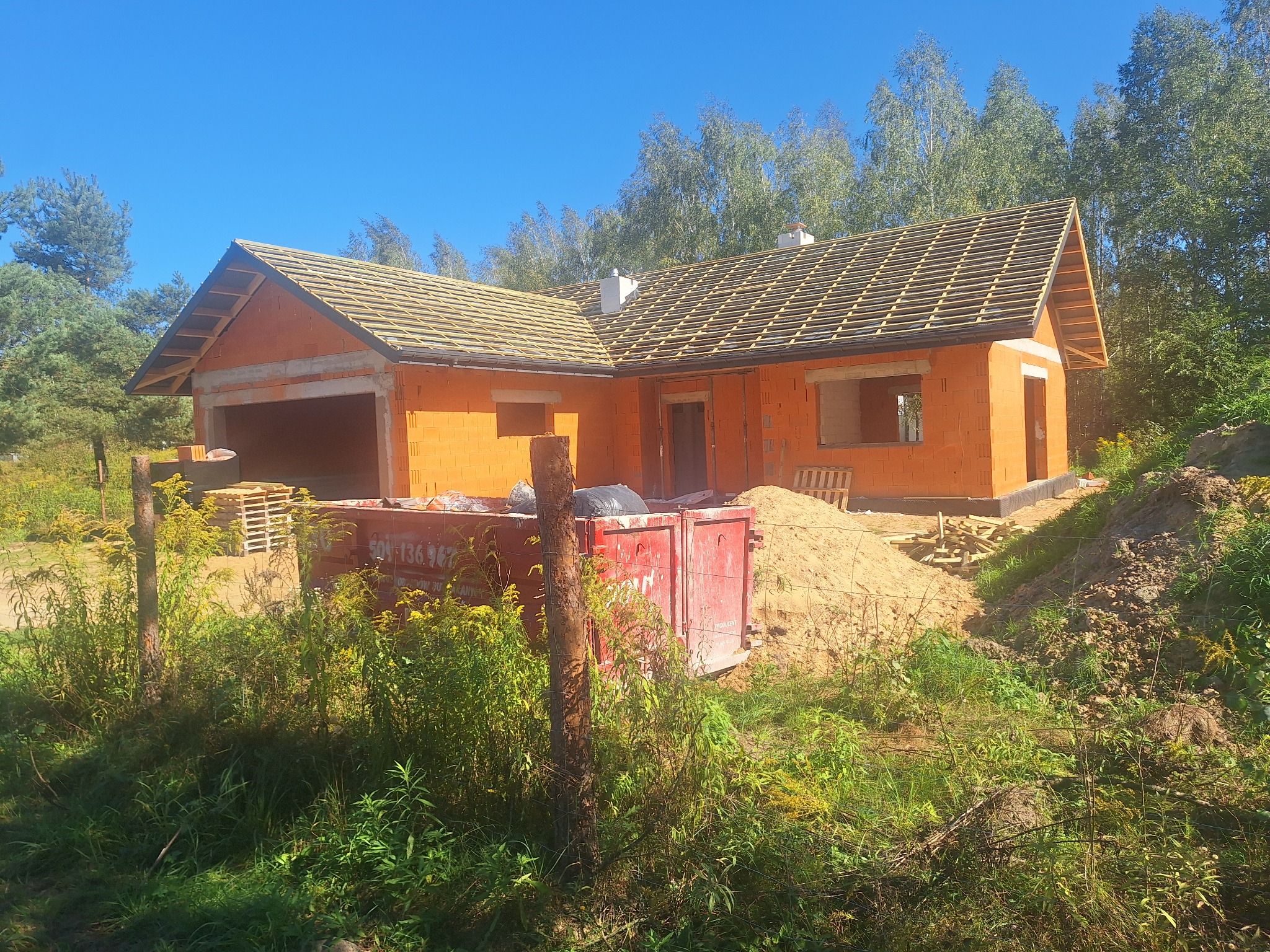 Surowy dom z pustaków z drewnianą konstrukcją dachu w trakcie budowy, widok z zewnątrz na tle zieleni i błękitnego nieba, kontener na gruz na pierwszym planie.