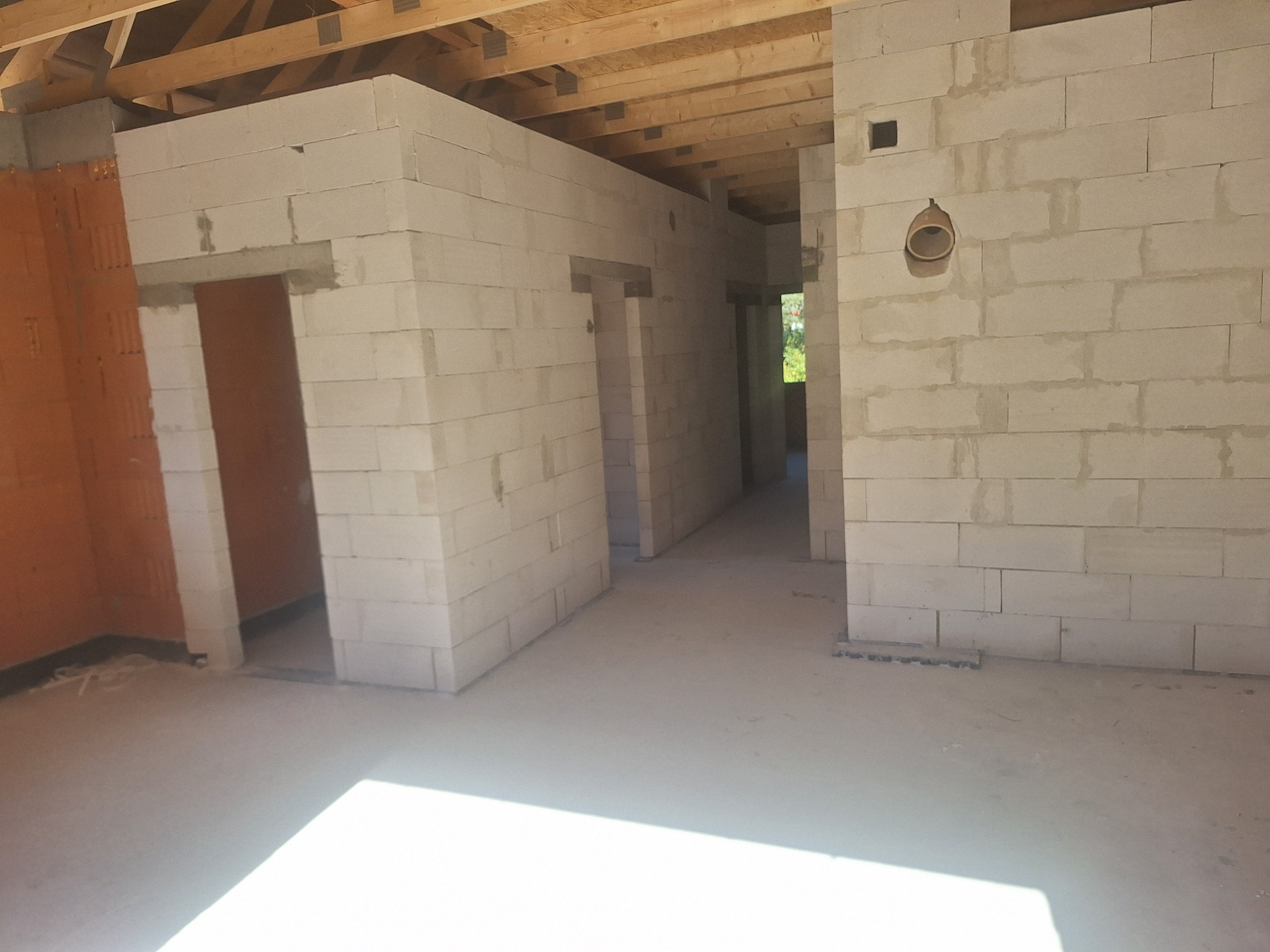 Surowy stan wnętrza domu w trakcie budowy: ściany z bloczków, widoczne otwory drzwiowe i drewniany strop. Brak wykończeń, surowy beton na podłodze.