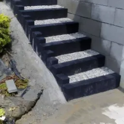 Nowe schody zewnętrzne z czarnych bloczków betonowych wypełnionych szarym grysem, przylegające do muru oporowego z szarego betonu.