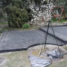 Układanie ciemnoszarej kostki brukowej na przygotowanym podłożu w ogrodzie, widoczne narzędzia i resztki materiałów budowlanych, w tle kwitnący krzew magnolii i huśtawka.