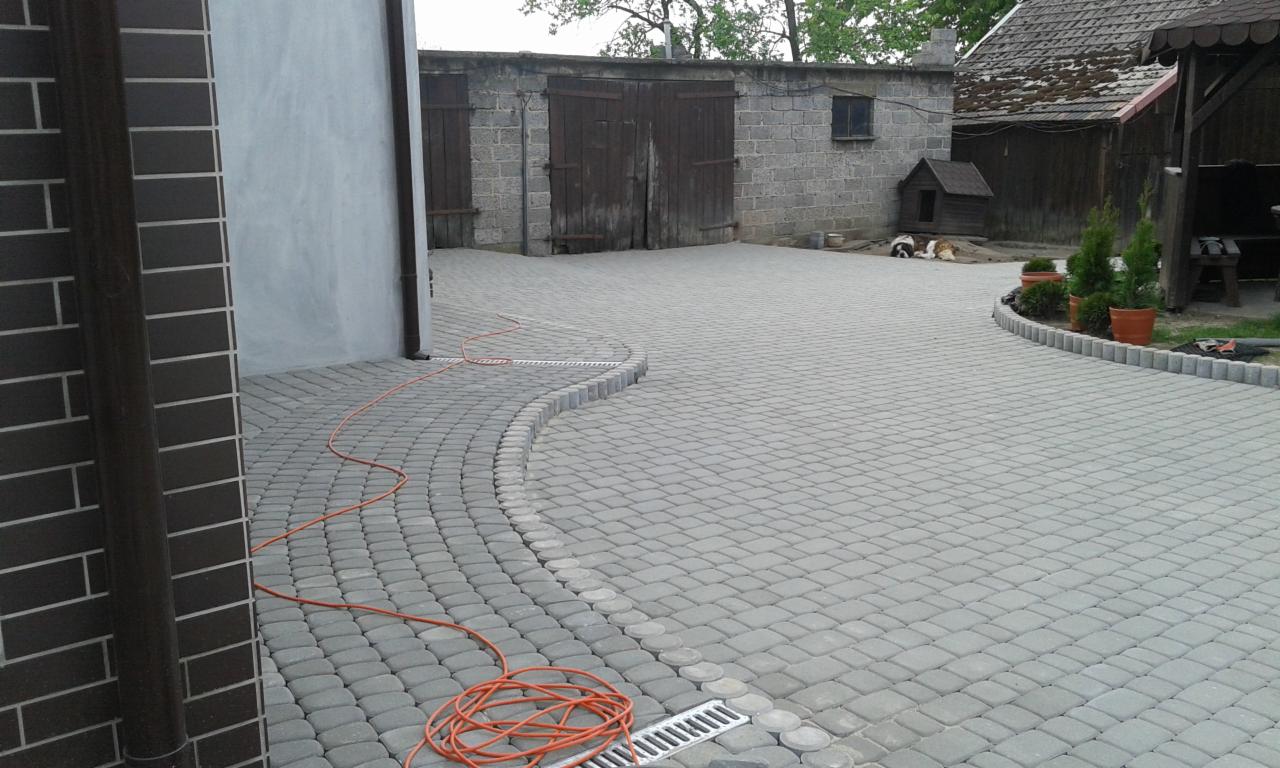 Szara kostka brukowa ułożona na posesji, z widocznym systemem odprowadzania wody i elementami małej architektury w tle. Widoczne dwa psy w budzie.