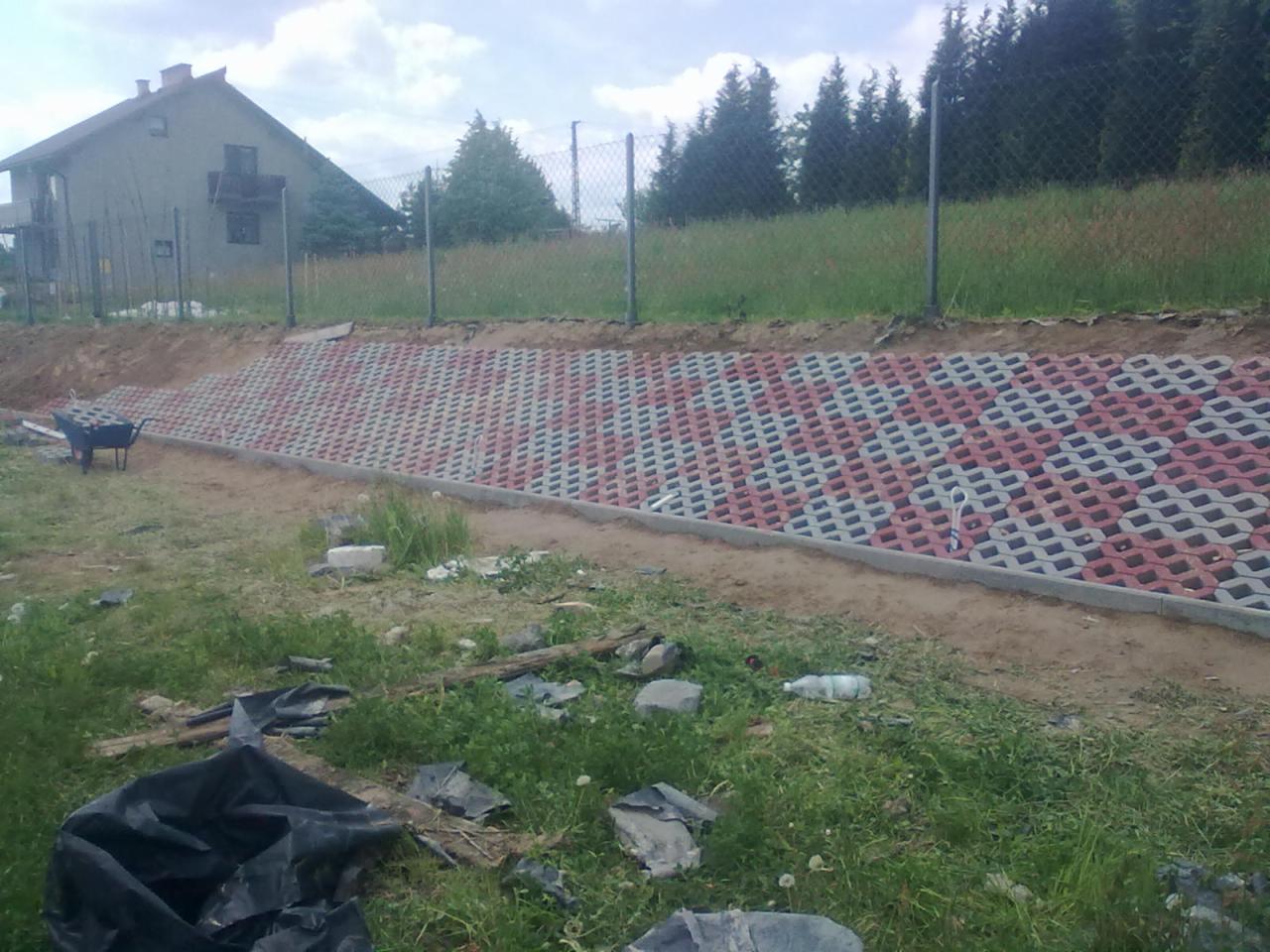 Skarpa umocniona ażurową kostką brukową w kolorach szaro-czerwonych, z widocznym domem w tle i częściowo rozplantowaną ziemią.