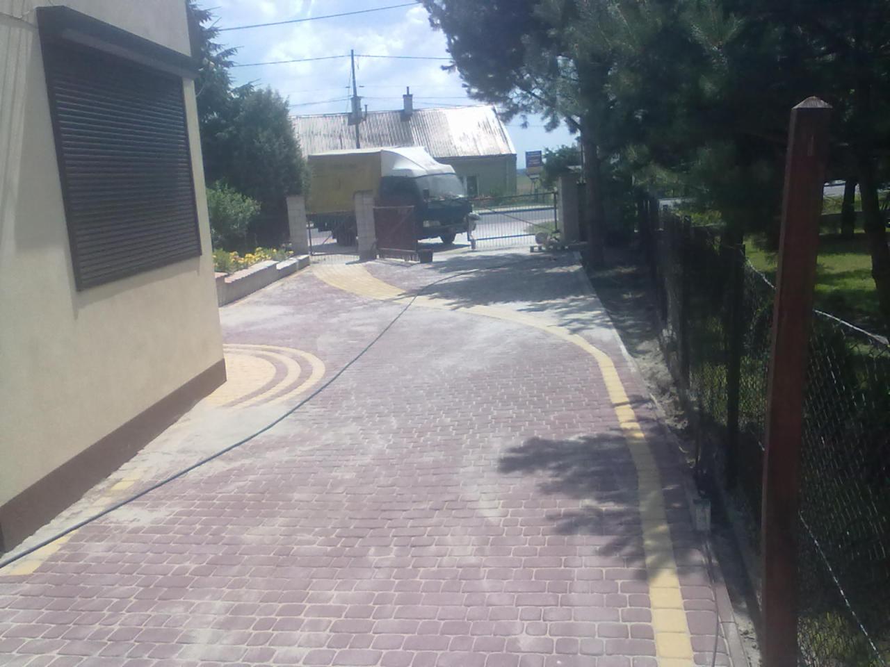 Czerwona kostka brukowa ułożona na podjeździe, z żółtymi akcentami i widocznym ogrodzeniem oraz budynkiem w tle. Na podjeździe stoi samochód dostawczy.