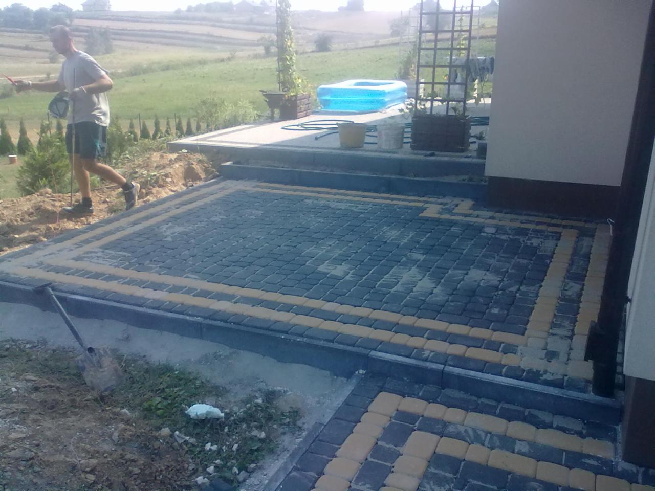 Ułożona kostka brukowa w geometryczny wzór z żółtymi akcentami, widok z góry, zarys budynku w tle, teren w trakcie prac wykończeniowych.