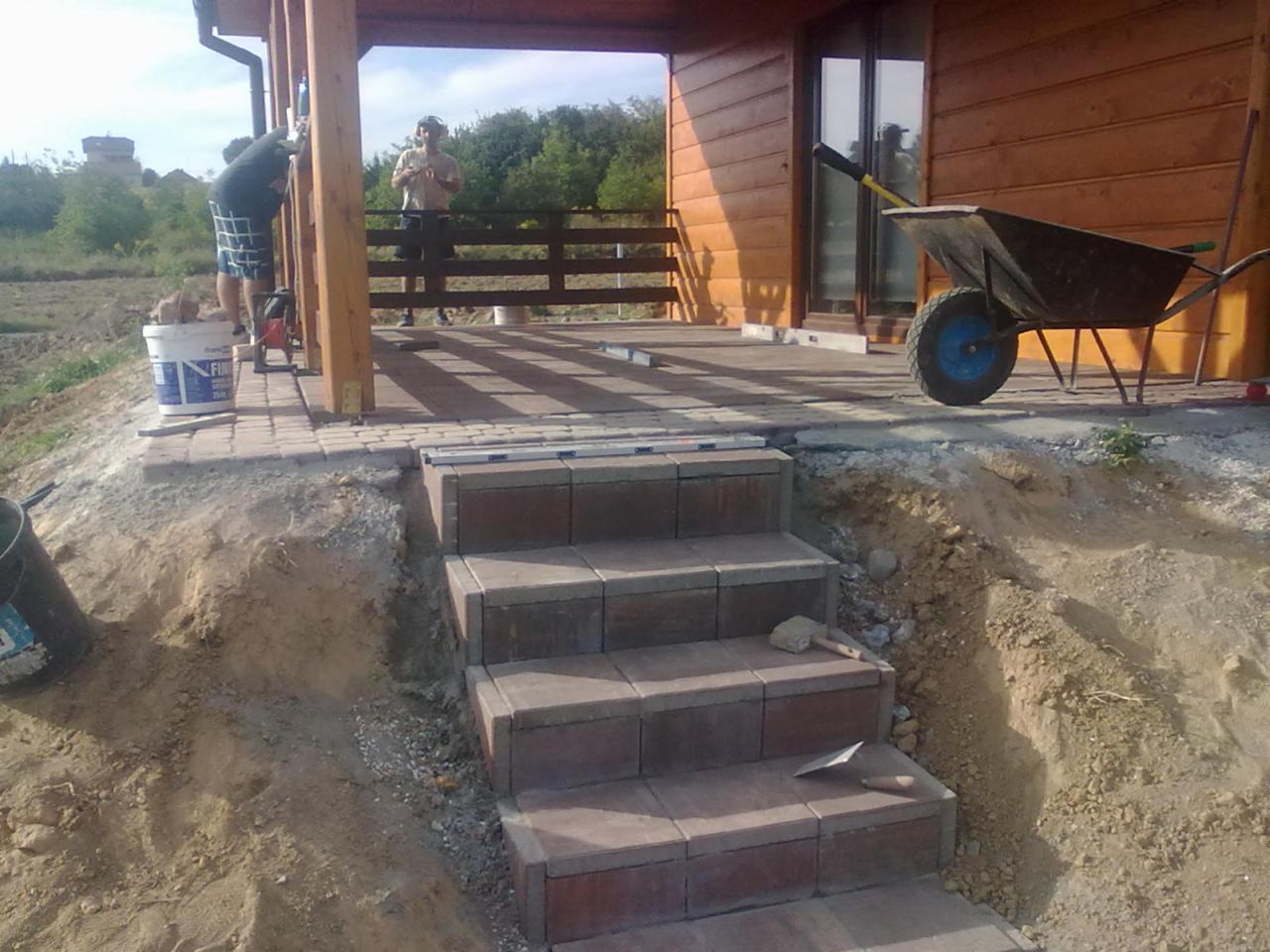 Budowa schodów z kostki brukowej prowadzących na taras drewnianego domu. Widoczna taczka i narzędzia brukarskie. Prace w toku, teren wokół w trakcie zagospodarowania.