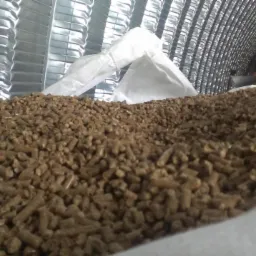 Jasnobrązowy pellet drzewny w worku, widok z bliska. W tle blaszane ściany magazynu z widocznymi elementami konstrukcyjnymi.