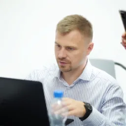 Mężczyzna w koszuli w paski skupiony na laptopie Dell, w tle osoba z telefonem i butelką wody.