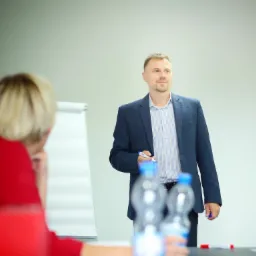 Prelegent w marynarce trzymający marker podczas prezentacji, w tle flipchart i słuchacz