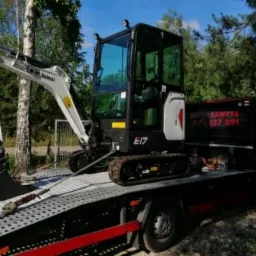 Mini koparka Bobcat E17 na lawecie, zabezpieczona linami, widoczny numer telefonu lawety na burcie pojazdu.