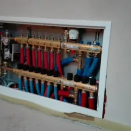 Rozdzielacz ogrzewania podłogowego TECE w białej wnęce ściennej, z widocznymi rurami w kolorach czerwonym i niebieskim, oraz elementami instalacji miedzianej.