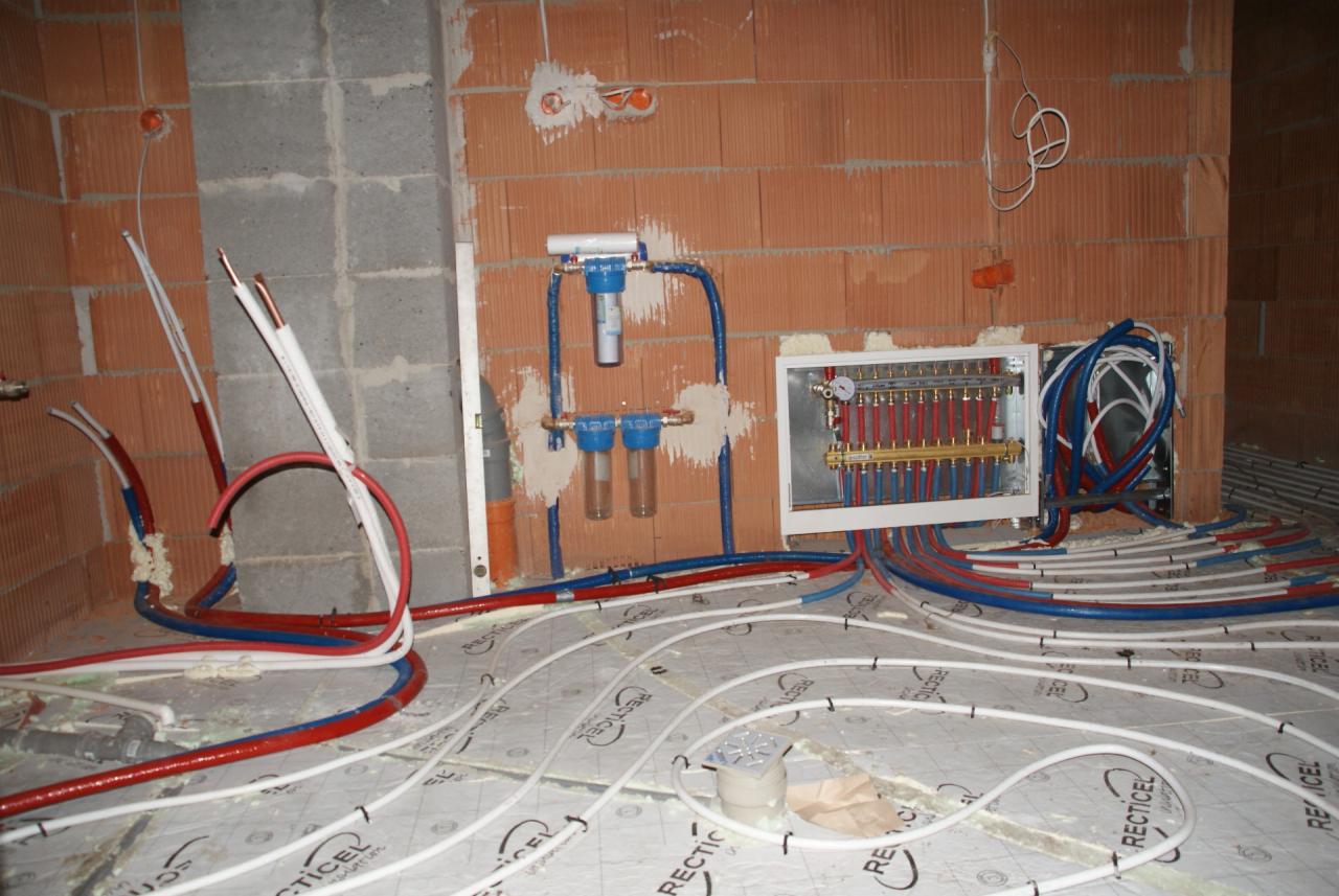 Instalacja ogrzewania podłogowego w trakcie montażu: rozłożone białe rury na izolacji, kolektor i filtry na ścianie z cegły. Widoczne kable elektryczne.