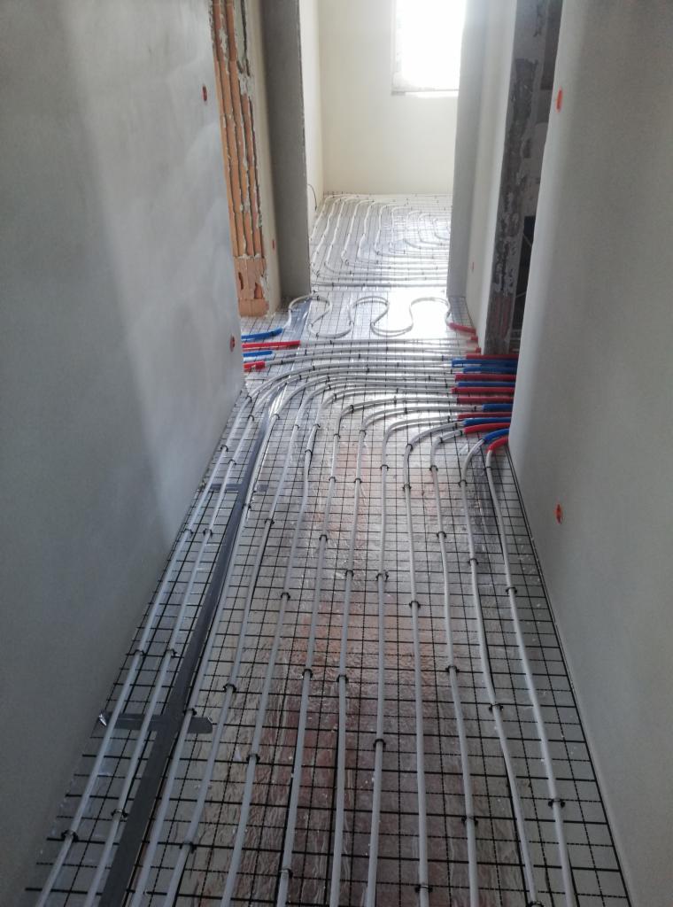 Montaż rur ogrzewania podłogowego na metalowej siatce w korytarzu, widoczne fragmenty ścian w stanie surowym i okno w oddali.