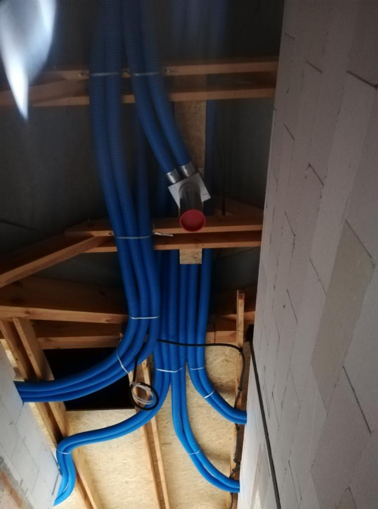 Niebieskie, karbowane rury wentylacyjne zamontowane pod drewnianym stropem poddasza, połączone z metalowym rozdzielaczem i zabezpieczone białymi opaskami zaciskowymi, widok z dołu.