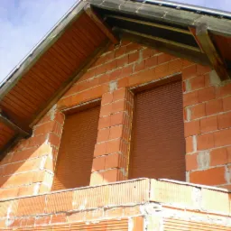 Dwa okna z brązowymi roletami w surowym murze z czerwonej cegły pod skośnym dachem, widok z dołu w słoneczny dzień.