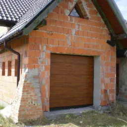 Brązowa brama garażowa segmentowa w surowym budynku z cegły. Widoczne niedokończone detale elewacji i brak tynku. Ujęcie z zewnątrz, w dziennym świetle.