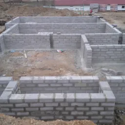 Wznoszenie ścian fundamentowych z bloczków betonowych na gruncie. Widoczny zarys przyszłego budynku. W tle palety z bloczkami i budynek magazynowy.