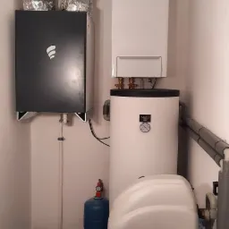 Kompletna instalacja grzewcza w kotłowni: czarny moduł z logo, biały bojler z manometrem, niebieski zbiornik wyrównawczy i srebrne rury wentylacyjne pod sufitem.