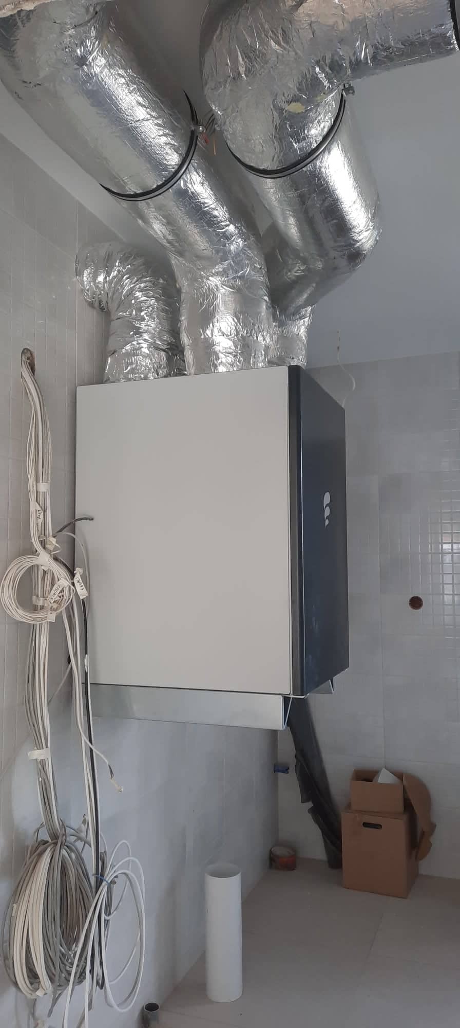 Rekuperator z izolowanymi przewodami wentylacyjnymi na suficie, widoczne kable elektryczne i niedokończone wykończenie wnętrza.