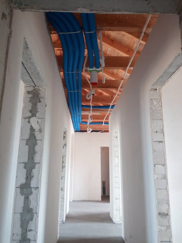 Wnętrze w trakcie prac instalacyjnych: widok korytarza z nieotynkowanymi ścianami, pod stropem ułożone niebieskie rury wentylacyjne przymocowane do drewnianej konstrukcji dachu.