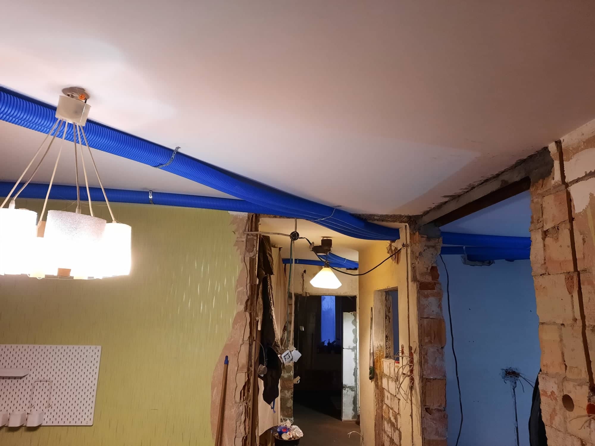 Niebieskie rury wentylacyjne poprowadzone pod sufitem w remontowanym mieszkaniu, widoczne fragmenty ścian z odsłoniętą cegłą i instalacjami elektrycznymi, w tle widoczne przejście do innego...