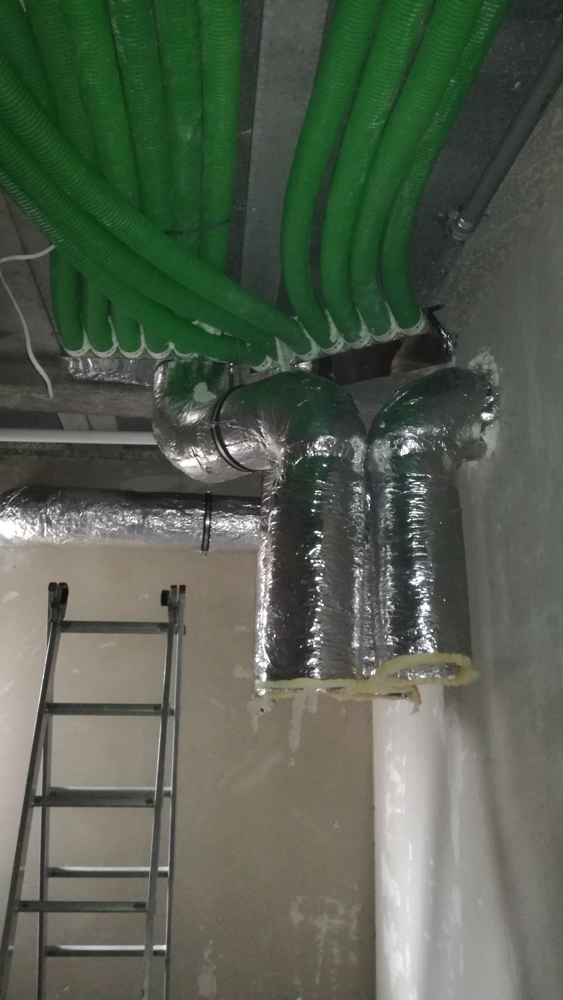 Zielone, karbowane rury wentylacyjne połączone z izolowanymi kanałami, widoczne pod sufitem w surowym wnętrzu, drabina oparta o ścianę.