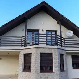 Dom jednorodzinny z brązowymi roletami zewnętrznymi, balkonem z metalową balustradą i anteną satelitarną na elewacji.