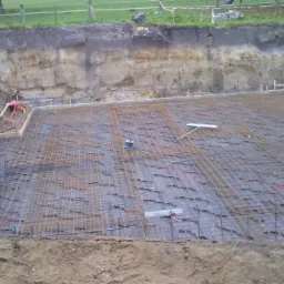 Siatka zbrojeniowa ułożona w wykopie przygotowanym pod fundament, widoczne szalunki i pracownik z maszyną do zagęszczania gruntu w oddali.