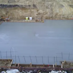 Świeża wylewka fundamentowa na placu budowy, zabezpieczona zbrojeniem i drewnianymi szalunkami, z widoczną drabiną i wykopem w tle.